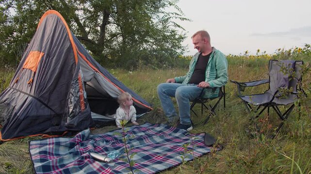 Outdoor parenting scene with family and baby near tent. Leisure activity on picnic blanket in grassy landscape