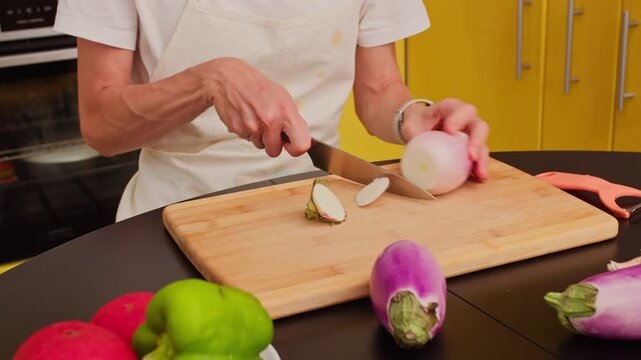 Person cuts the stem from a fresh purple eggplant on a wooden board amidst other vegetables
