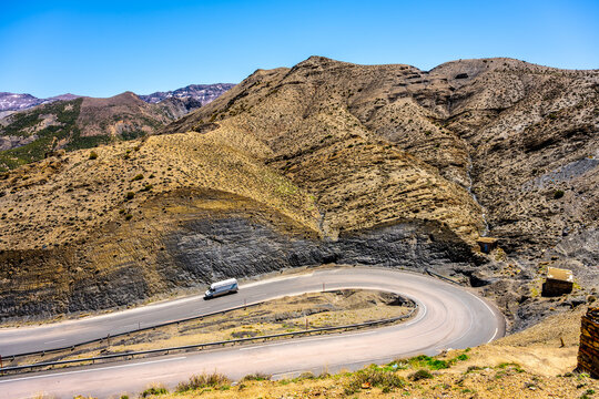 A road curves around the hills at Tizi-n-Tichka Pass. The scene shows dry mountains under a clear sky. Travelers can be seen driving along the winding path.
