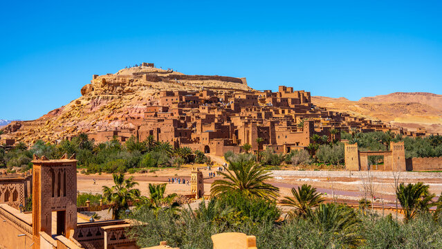 People explore Ait Benhaddou, a historic ksar in Morocco. The village shows traditional earthen clay architecture. It is set against desert hills under a blue sky.