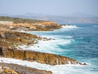 Waves crash against the rocky coast of the Atlantic Ocean in southern Morocco. The landscape shows cliffs and distant mountains under a bright sky. © pyty