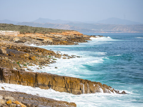 Waves crash against the rocky coast of the Atlantic Ocean in southern Morocco. The landscape shows cliffs and distant mountains under a bright sky.