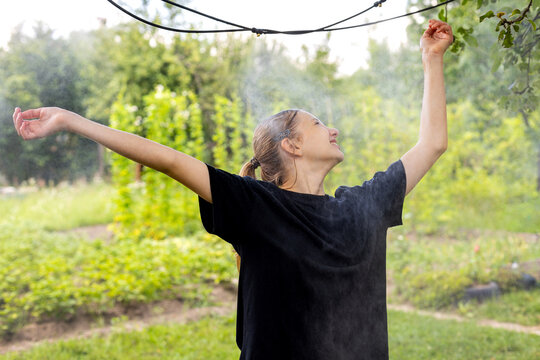 Young Woman Enjoying Cooling Garden Mist Outdoors. Young woman smiles with arms raised while enjoying a fine cooling mist in a lush summer garden.