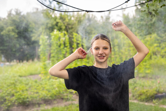 Teenage Girl Enjoying Cooling Water Mist in a Garden. Teenage girl in a black shirt raises her arms beneath a cooling water mist in a lush green garden.