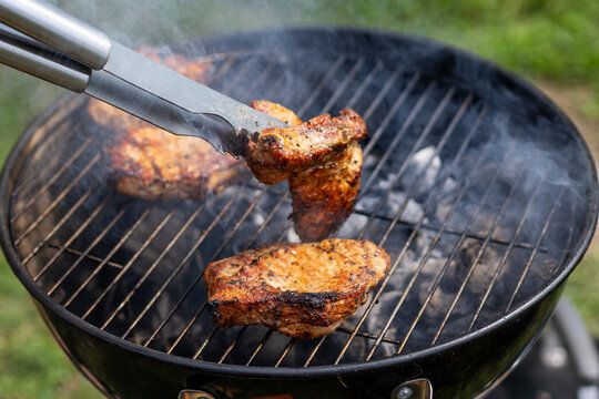 Tongs Lifting Grilled Pork Chop Over Smoky Charcoal Barbecue. Metal tongs lift a browned pork chop above a smoky charcoal grill while seasoned cuts cook over glowing coals outdoors.
