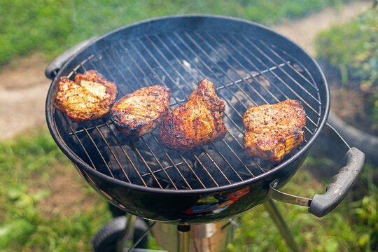Four Pork Chops Cooking on a Smoky Charcoal Grill. Four seasoned pork chops cook on a smoky round charcoal grill outdoors with browned surfaces and dark.
