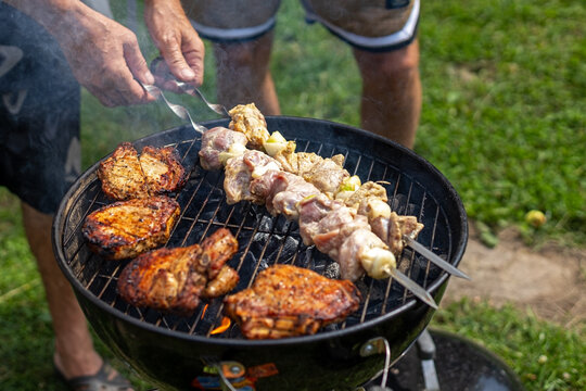 Person Grilling Pork Chops and Meat Skewers Over Charcoal. A person turns marinated meat and onion skewers beside browned pork chops on a smoky charcoal grill outdoors.