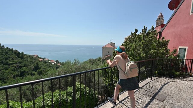 Cervo, liguria, italy. June 22, 2025. Female tourist with a backpack admiring the stunning panoramic view of the mediterranean sea from a balcony in the historic village of cervo