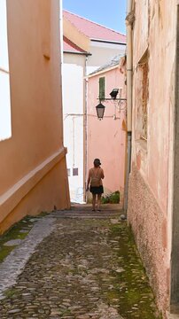 Cervo, liguria, italy. June 22, 2025. Rear view of a young woman with a backpack walking down a picturesque narrow cobblestone alley in the historic medieval village of cervo, liguria