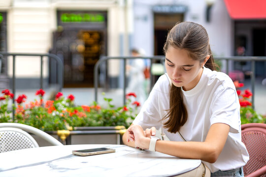 Young Woman Checking Smartwatch at Outdoor Cafe. Young woman checks a smartwatch while seated at an outdoor cafe table with a smartphone nearby and colorful flowers in the background.