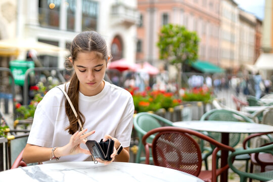 Young Woman Opening Wallet at Outdoor City Cafe. Young woman opens a black wallet while seated at an outdoor cafe table in a busy city square.