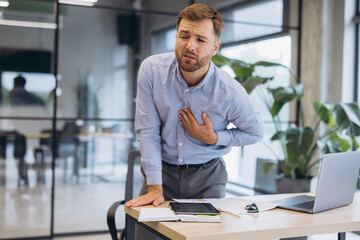 Obraz premium Businessman feeling severe chest pain, clutching his heart, experiencing stress and illness in a modern office environment
