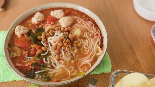 A wholesome homemade meal featuring a bowl of spicy noodle soup with vegetables, fish balls, chickpeas, and rice noodles, served alongside fresh avocado halves and sliced apples.