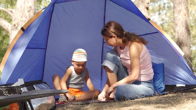 A happy mother and son sit together inside their blue tent, playing quietly. They feel content and bonded while relaxing in the sunny pine forest.