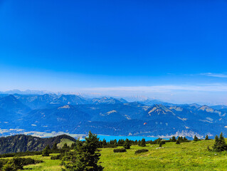 Obraz premium A vast mountain landscape unfolds from a grassy green peak, showing a bright blue lake in the valley below and a distant paraglider.