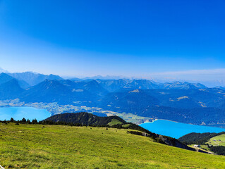 Fototapeta premium A picturesque panoramic view shows a vibrant turquoise lake nestled between rolling blue mountains under a clear sky, captured from a grassy alpine meadow.