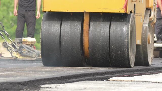Yellow road roller compacts fresh asphalt. Large black drums press the surface firmly. Light smoke rises from the drums. Worker in black pants stands nearby. Green landscape shows outdoor setting