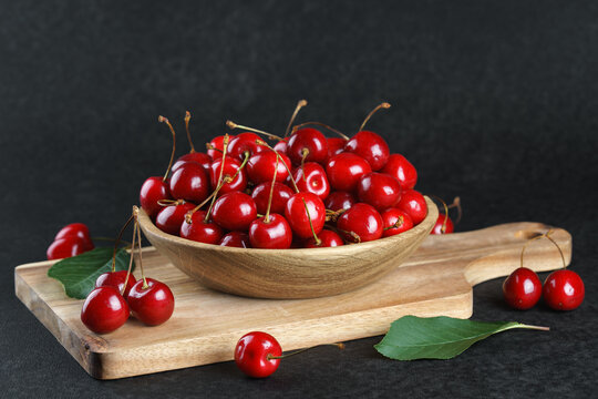 Fresh ripe sweet cherries in wooden bowl on rustic cutting board with green leaves and linen cloth on black textured background. Healthy summer fruit, red berries, fresh seasonal harvest, healthy food