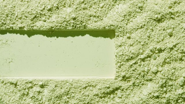Above view of pale green natural powder surface showing a stretched rectangular pattern lit by orbiting light casting dynamic shadows | Green clay cosmetic ingredient concept