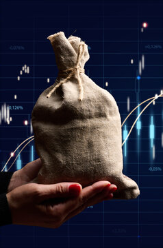 A woman's hands hold a tied canvas bag of money against a blurred digital stock market chart