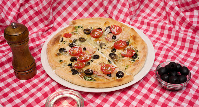 Pizza with cheese, tomatoes and olives on a white plate standing on a checkered red tablecloth