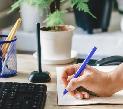 A hand is writing something on paper at a wooden desk. A live houseplant stands in a white pot nearby. A keyboard and pen holder are visible on the surface. The environment is conducive to calm and pr