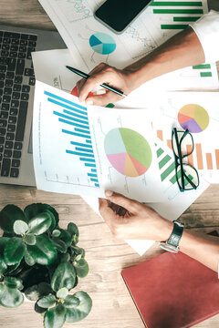 Person analyzes financial charts with focused attention. Hand holds graph while pen rests nearby. Green plant adds calm to workspace. Laptop and scattered papers suggest active review