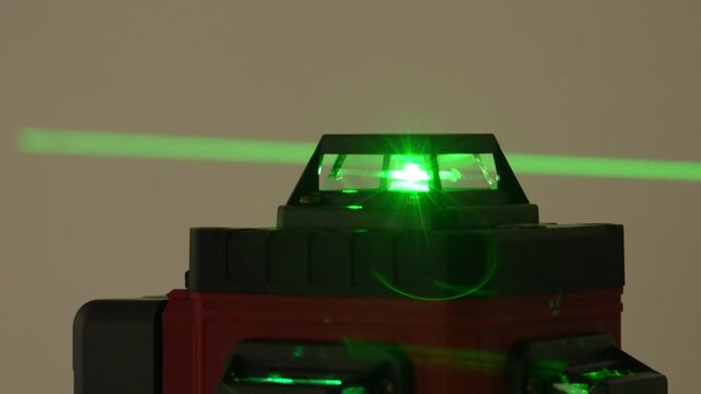 Single green laser beam projects horizontally across a beige background. Red and black laser pointer emits the steady beam. Bright light highlights precision measurement technology