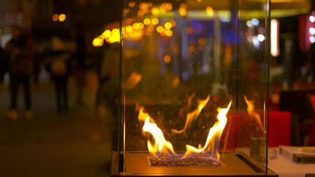 Decorative glass fireplace burning on an outdoor cafe terrace at night. Modern portable fireplace with a burning flame providing warmth and ambiance on a restaurant patio, with people blurred