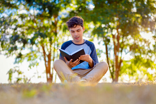 Brazilian man reading a book outdoors in a green park during golden hour, representing education, leisure, concentration, personal growth, and the joy of reading in nature.