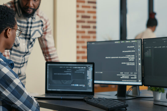 Software developer writes code on a laptop while a coworker asks for advice, showing programming teamwork and cloud computing collaboration.