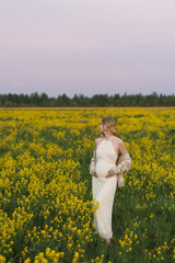 Obraz premium Pregnant woman stands among dense yellow flowers in wide meadow under soft pastel sky, wearing light dress and cardigan. Calm outdoor portrait with gentle spring colors and serene rural mood