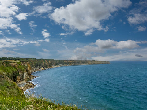 anoramic view of the steep limestone cliffs at Pointe du Hoc in Normandy, France, overlooking the blue waters of the English Channel under a cloudy sky. A historic World War II