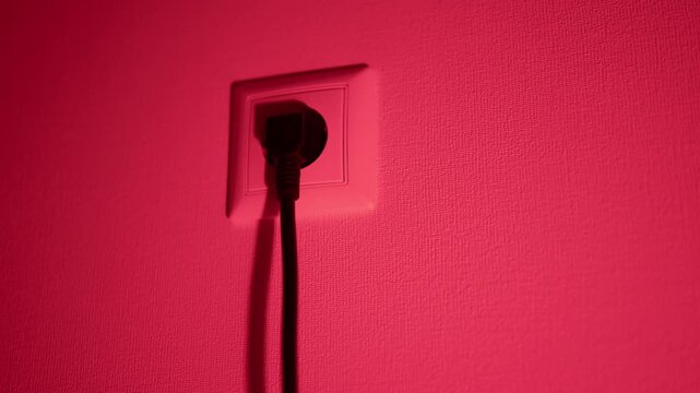 Black power plug in wall socket on textured pink illuminated wall with dramatic shadow