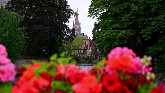 Slow-motion panoramic view of a Bruges canal with a historic church, stone bridge, tourists, and colorful flowers. Charming medieval cityscape in Belgium.