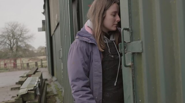 Young woman farmer in work overalls sliding a heavy corrugated metal barn door shut on a farm