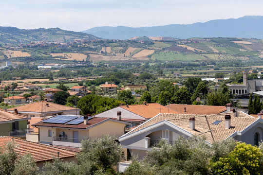 Panoramic view of residential rooftops with solar panels overlooking green agricultural fields and scenic rolling hills under bright sky in sunny countryside Italy.