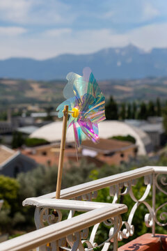 Colorful iridescent plastic toy pinwheel attached to white balcony railing overlooking scenic town and distant mountains on a sunny summer day in countryside Abruzzo Italy.