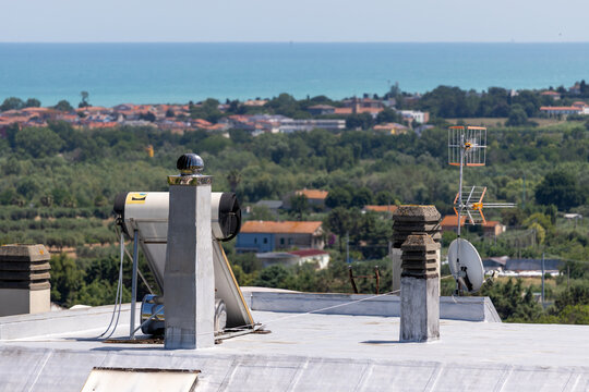 A solar water heating system on the roof of a house with chimneys and an antenna, overlooking the coastal landscape of a town and the blue Adriatic Sea under a sunny sky in Italy.