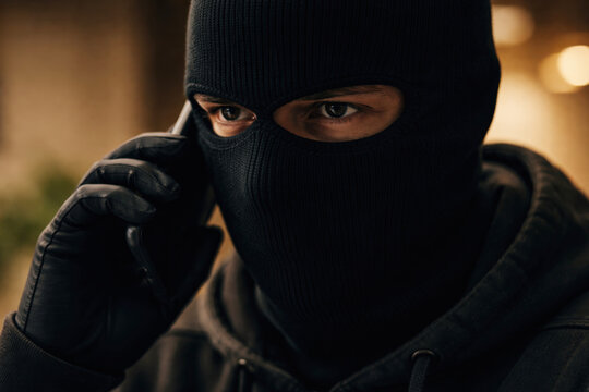 A person in a black balaclava and gloves makes a secretive phone call. This image is ideal for illustrating cybercrime, fraud, security breaches, and criminal activities.