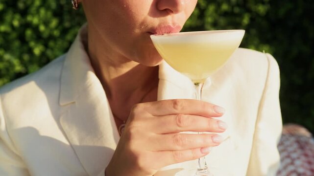 Close-up woman sipping pale yellow cocktail outdoors in warm sunlight with elegant cream blazer
