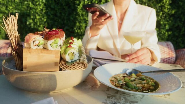 Elegant woman checking smartphone during outdoor sushi dinner with cocktail and fresh plates