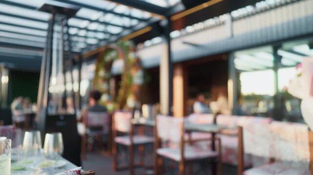 Blurred modern restaurant terrace with sunlit dining tables, glassware, chairs, and guests in warm evening light