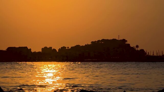 Golden hour timelapse of Denia Castle silhouette over Mediterranean Sea with calm water reflection, Spain
