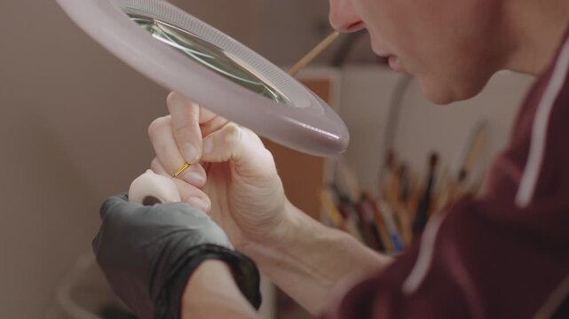 Artist painting facial details on a female PLA ball jointed doll under a magnifying lamp in a professional doll customization workshop.