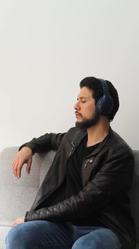 Vertical detailed view of a man wearing a leather jacket sitting on a grey couch listening music on a headset in tranquil attitude
