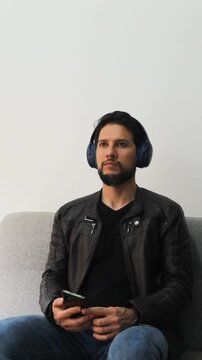 vertical detailed view of a smiling latin man listen music on a wireless headset and a smart phone sitting on a couch