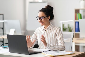 Fototapeta premium Smiling Young Mixed Race Woman Engaging In Video Call While Working At Home Office. Sitting At Desk With Laptop, Enjoying Professional Interaction In Modern Workspace. Business Concept.