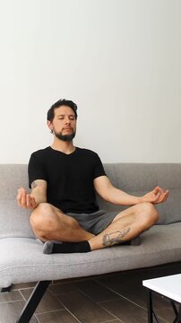 vertical detailed view of a man sitting at the left side of a couch in the lotus pose meditating