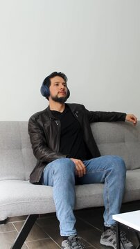 Vertical view of a latin man in a relaxing attitude sitting on a grey couch listening music on a wireless headset